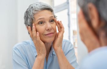 A concerned mature woman looking at her eyes and eyelids in a mirror.